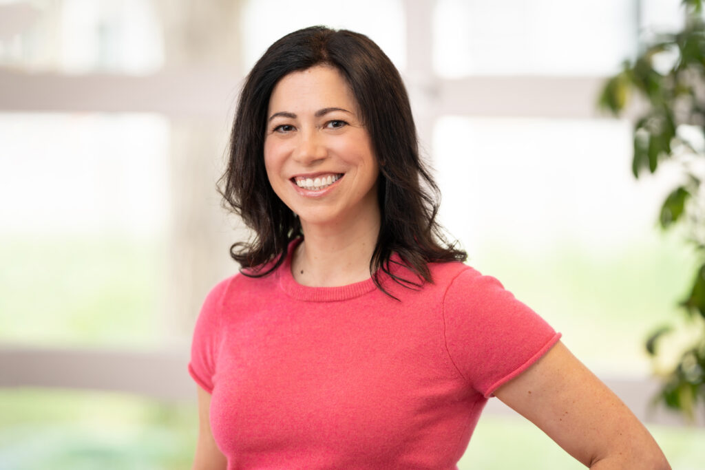 Headshot of Megan Vogels; a white woman with medium-length dark-brown hair and wearing a pink sweater; smiling and standing in front of a window. 