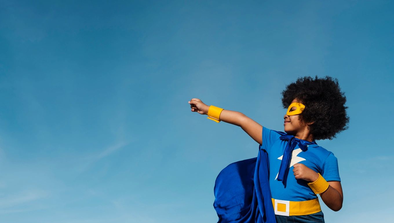A Black child wearing a superhero costume and mask, smiling and standing with a fist raised against a blue sky background.