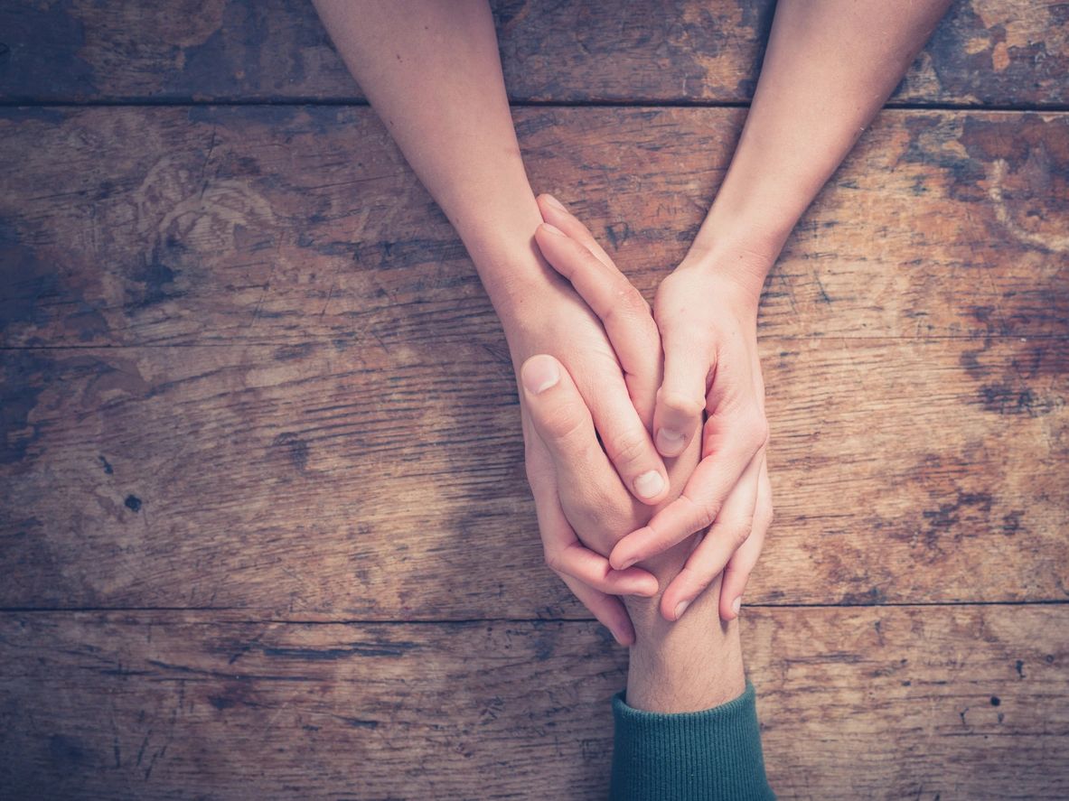 An overhead picture of two light-skinned hands clasping another light-skinned hand on a wooden table. 