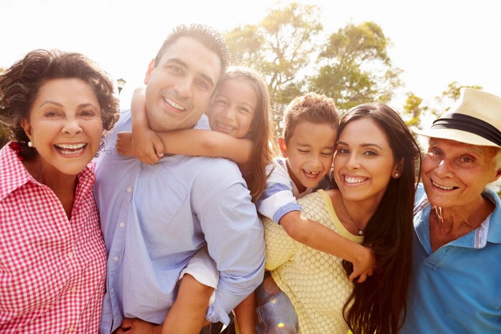 A happy family stands together outside with trees in the background. The two children are each on their parents' back, and a grandmother and grandfather stand on each side. All people are smiling and wearing bright colored shirts. 