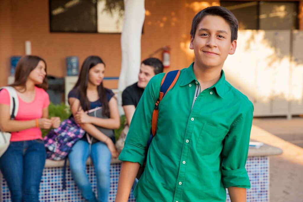 A smiling boy with dark brown hair is standing with a backpack over one shoulder. Three other students, 2 girls and a boy, are in the background.