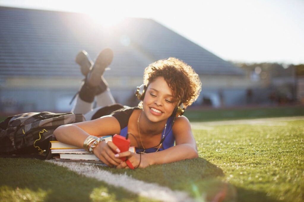 A smiling young person, with medium-colored skin and brown hair, lays on her stomach on a field, looking at her phone with her backpack and books beside her