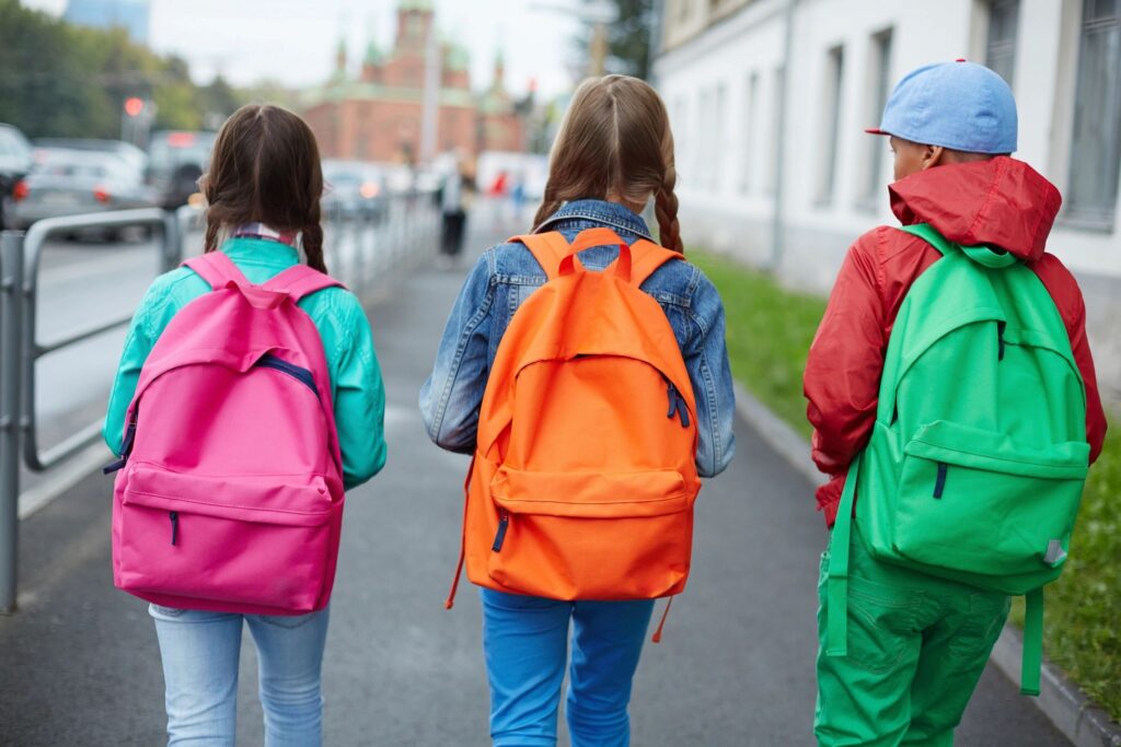 Three elementary school-aged children, two girls and a boy, are walking away, wearing jackets and brightly colored backpacks. 