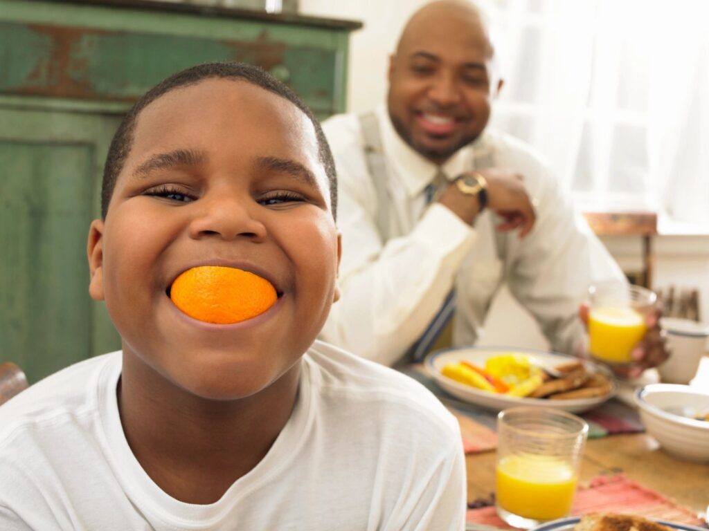 A black child faces the camera and smiles, with an orange in his mouth; A black man sits behind him at a table with breakfast food
