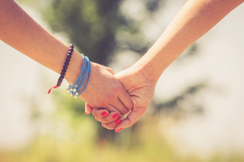 Two people hold hands outdoors; one person has pink nail polish and the other is wearing several colored bracelets