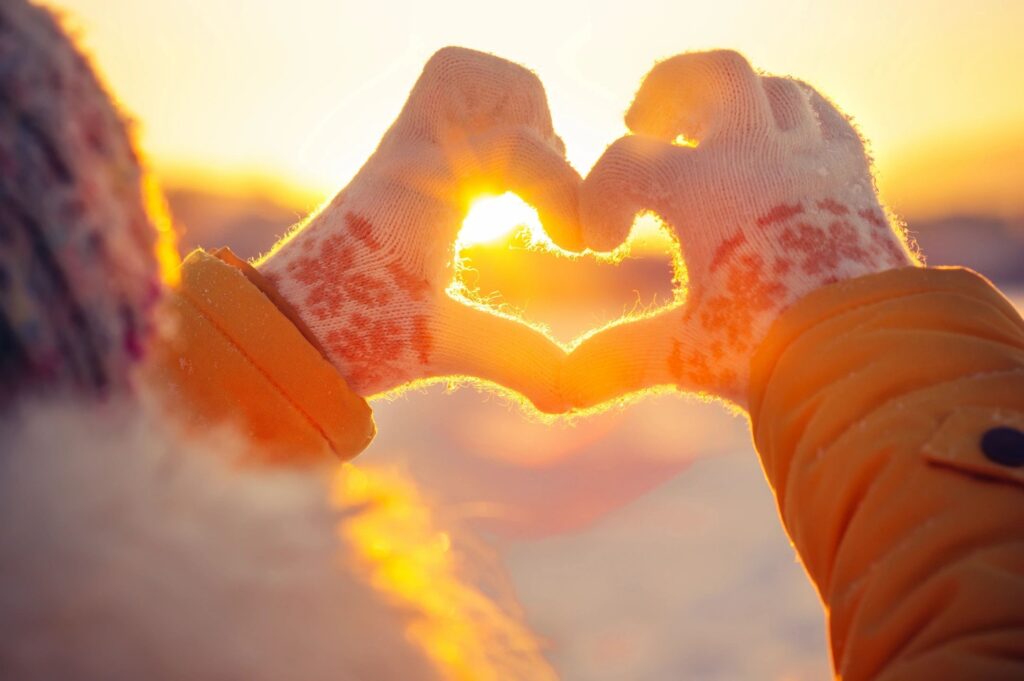 A person wearing mittens holds their hands in the shape of a heart in front of a setting sun