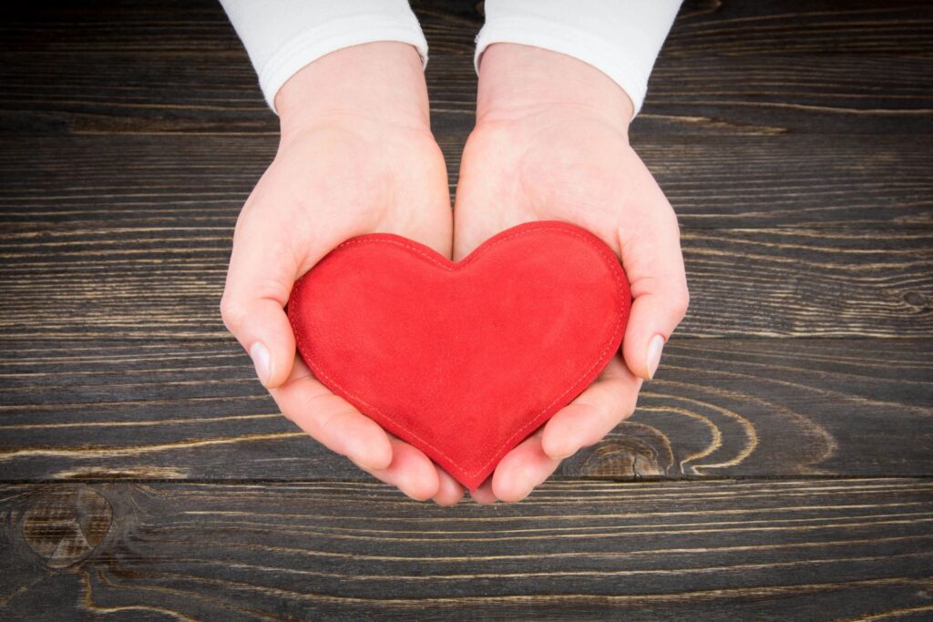 A person with light skin is holding a red heart in their hands against a wooden table