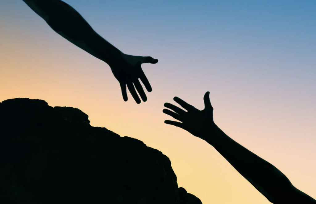 one hand reaching toward another against a sunset sky