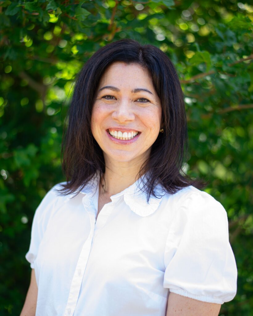 A headshot of Megan Vogels smiling, standing agains green trees